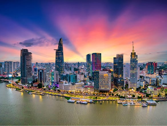 Panoramic view of a modern city skyline at sunset along a river, with tall glass buildings and boats along the shore.