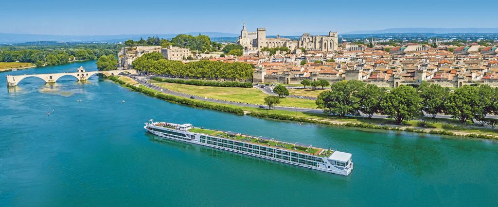 A large white river cruise boat glides along a turquoise river past a historic town with red-roofed buildings and a stone bridge, green trees lining the shoreline.
