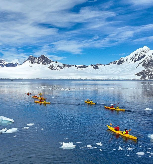 Yellow kayakers paddling across icy blue water with snow-covered mountains under a blue sky.