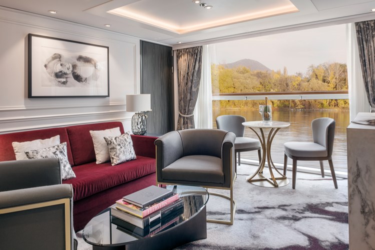 Stylish living room with a red sofa, gray chairs, and a glass coffee table, overlooking a lakeside view through large windows at dusk or daytime light.