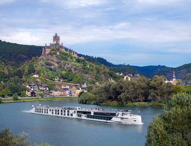 White river cruise boat glides on a calm river, with a forested hillside and a medieval castle perched atop a hill in the background.