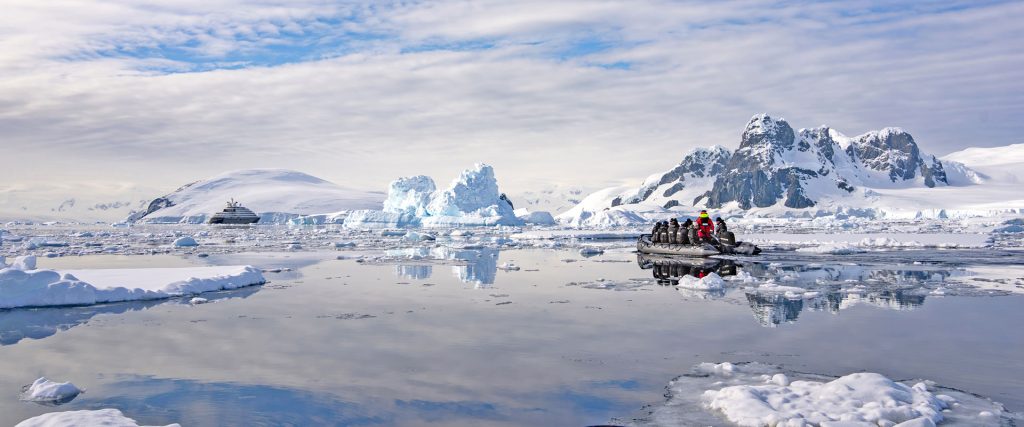 Zodiac inflatable boat with passengers and a guide navigating icy, reflective Arctic waters near jagged snow-covered mountains and icebergs at sunrise/sunset.