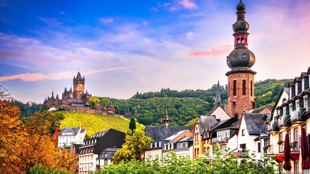 Picturesque European village with a tall church tower, colorful houses, and a castle on a hill amid autumn foliage.