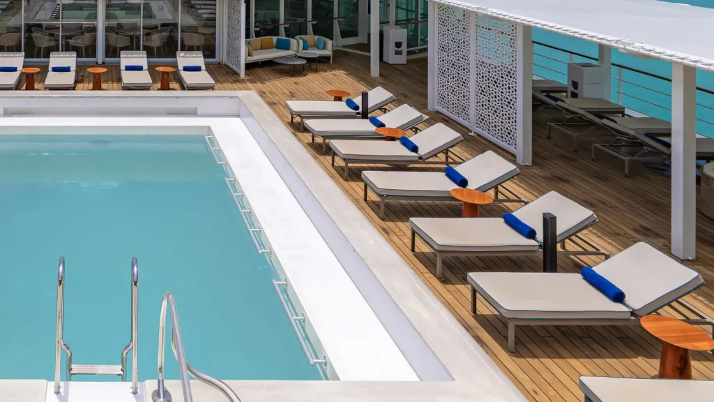 Poolside deck with white sun loungers, blue towels, and round wooden tables beside a turquoise pool on a wooden deck.