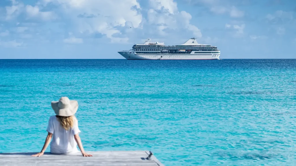 Person on a wooden dock gazing at bright turquoise sea with a large cruise ship on the horizon