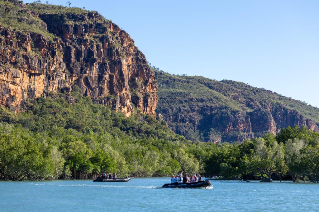 Two inflatable boats with people ride across a calm blue river, surrounded by green mangroves and a steep rocky cliff in the background.