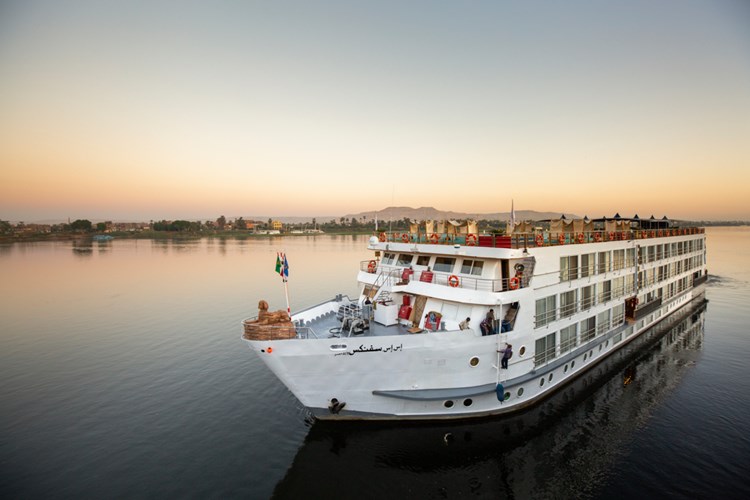 White multi-deck river cruise ship on calm water at sunset, passengers on the upper deck.