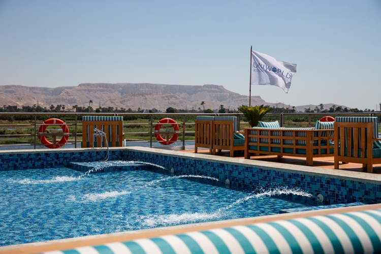 Outdoor pool area with blue mosaic tiles, wooden lounge chairs, striped cushions, red life rings, and a white flag against a desert landscape.