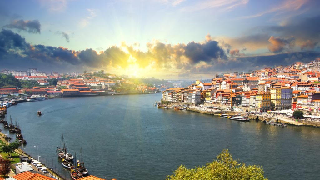 Sun rays over a riverfront city with colorful terraced buildings along the quay and boats on the water at sunset.
