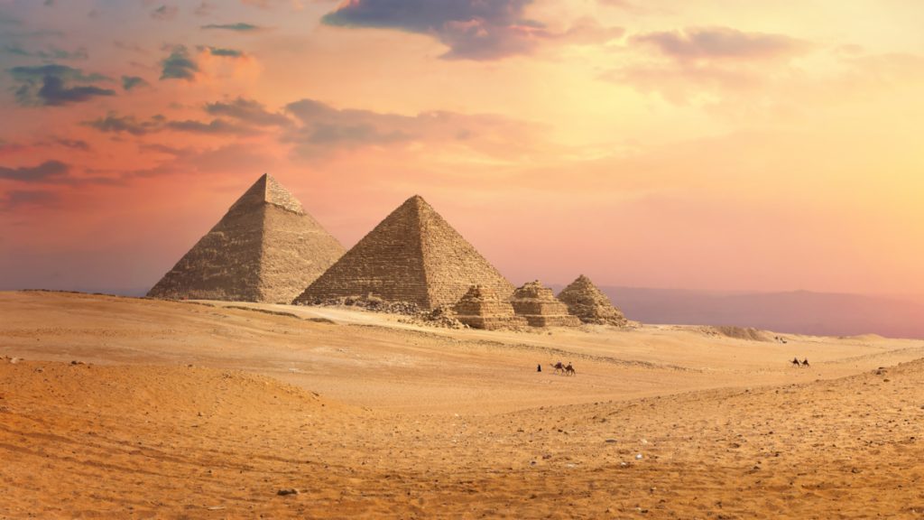 Pyramids of Giza at sunset, sandy desert foreground, small group of people on camels in the distance.