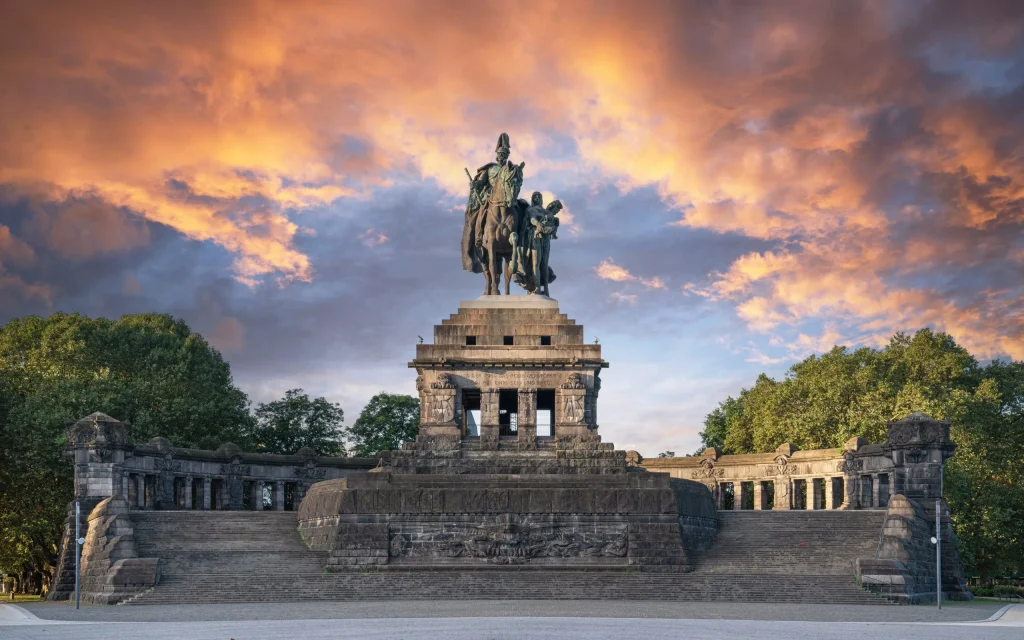 Equestrian statue on a multi-tiered stone pedestal, with a rider on horseback and a companion figure, framed by trees at sunset.