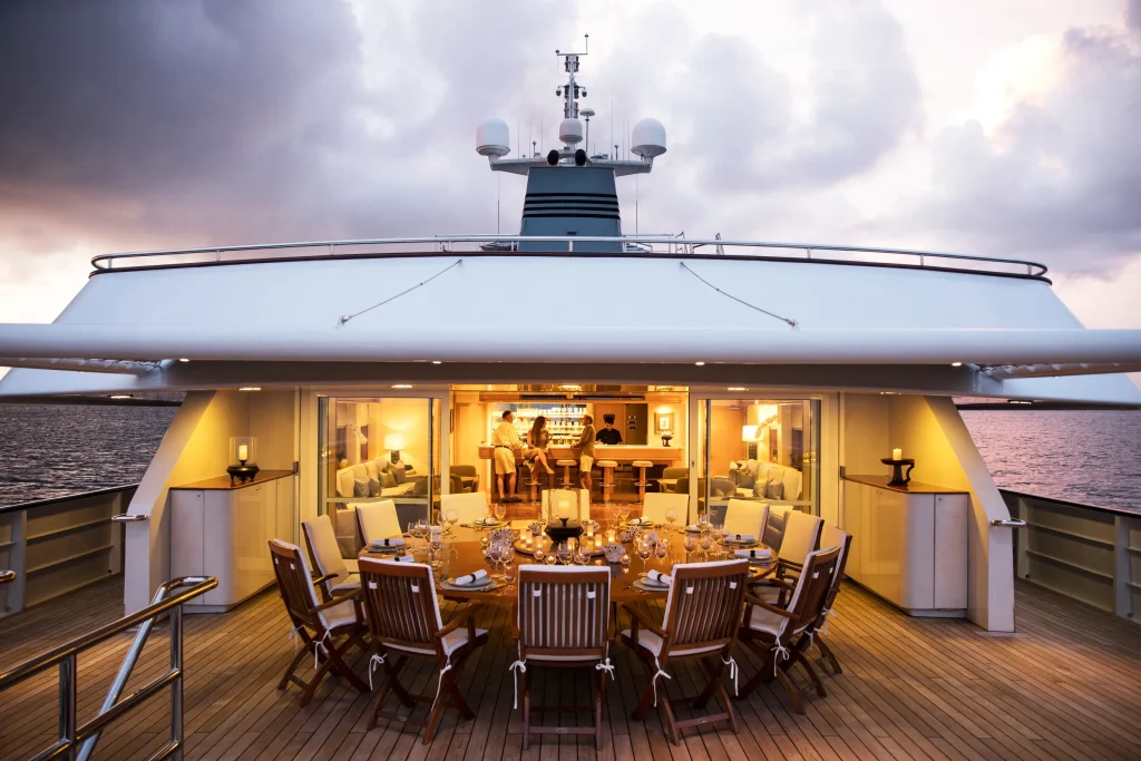 Open-air dining deck of a luxury yacht at sunset, round tables set with glassware and wooden chairs around a central table.