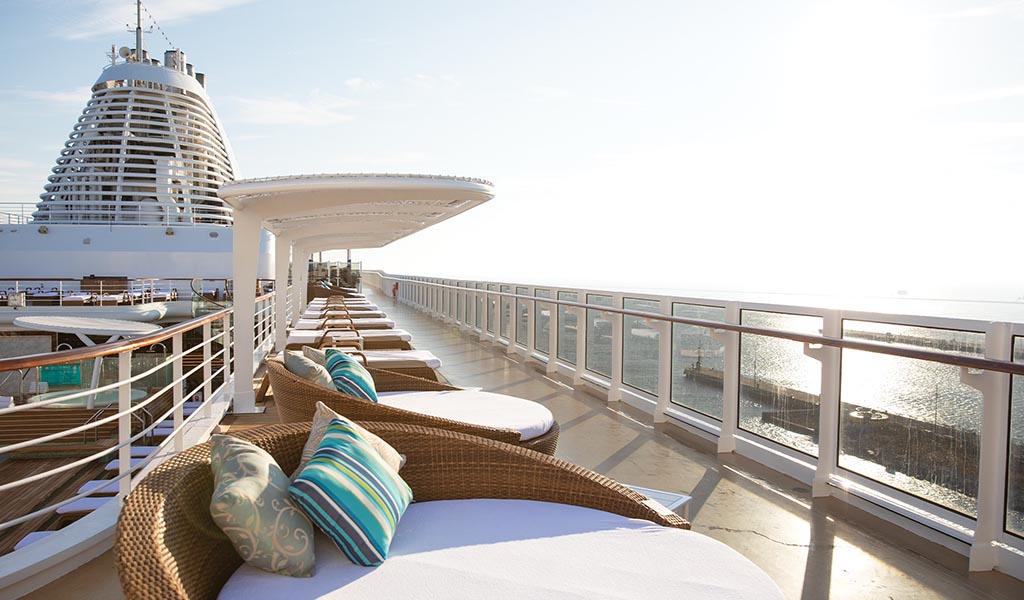 Sunlit cruise ship deck with wicker lounge chairs and striped cushions overlooking the ocean.