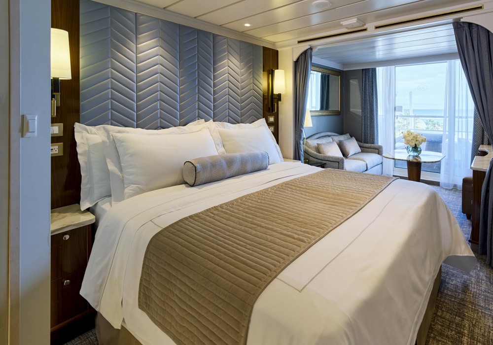 Hotel room with a large, neatly made bed featuring white linens and taupe bed runner; seating area by a balcony with ocean view.