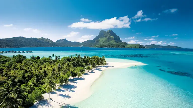 Tropical beach with white sand, palm trees, and turquoise waters against a blue sky and distant mountains