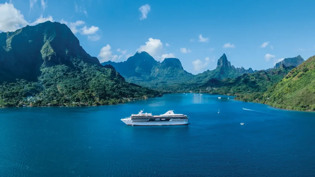 White cruise ship anchored in a tropical blue bay surrounded by forested mountains.