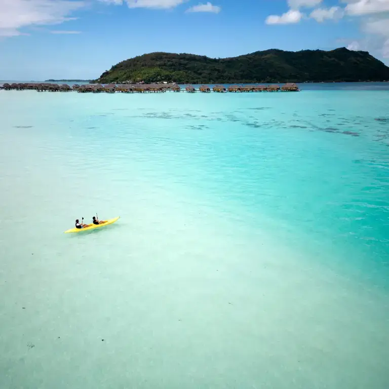 Two people paddling a yellow kayak across crystal‑clear turquoise water toward an island with overwater bungalows built on stilts along the shore