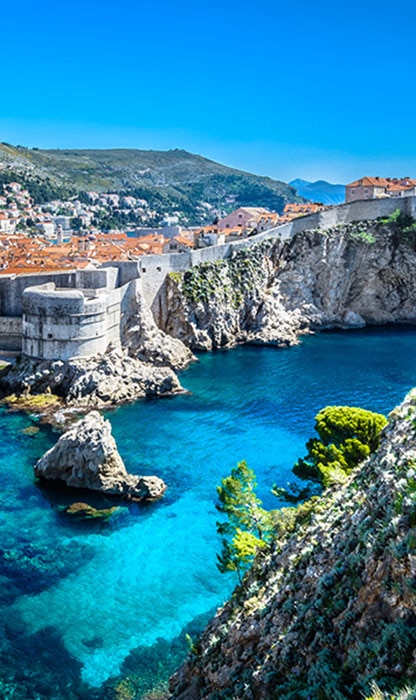 Coastal town built along rocky cliffs with orange-roofed houses, a stone fortress, and clear blue-green water offshore.