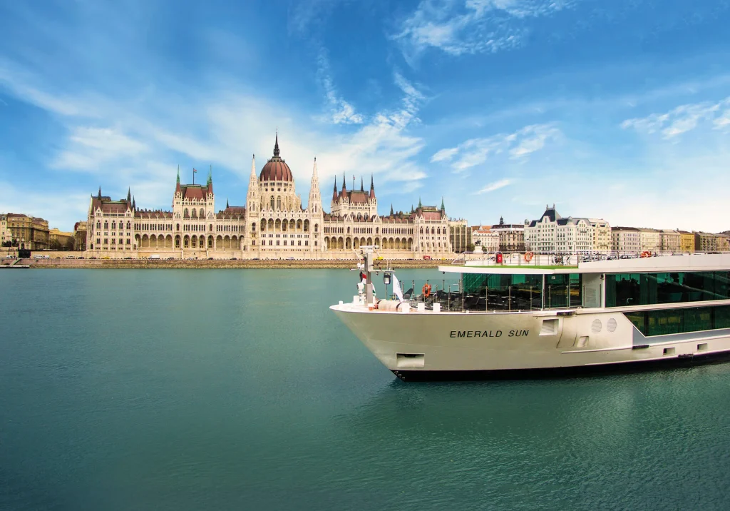 Budapest's Parliament building along the Danube with a large white cruise ship moored in front, under a blue sky.