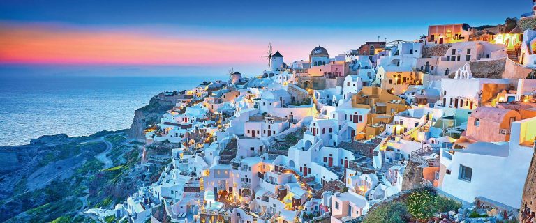 Sunset over a cliffside village of white-washed houses in Santorini, Greece, with warm lights and a blue sea liner in the distance.