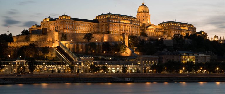 Buda Castle and fortress lit at dusk along the Danube River, Budapest, Hungary.