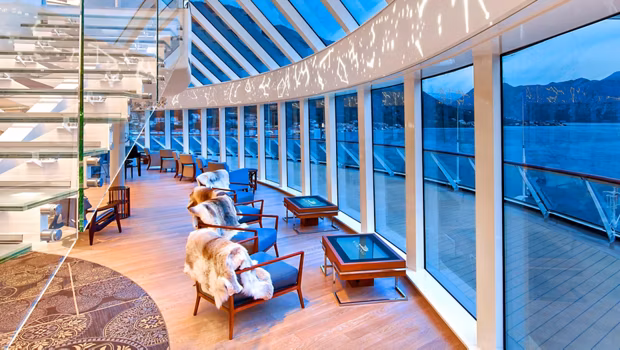 Luxurious glass-walled lounge with fur-covered chairs and glass tables, overlooking a blue ocean and mountains outside.