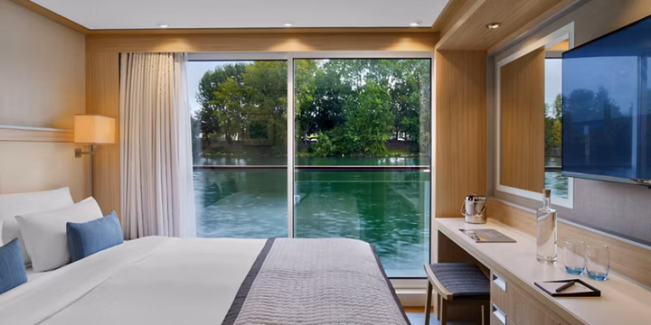 Hotel room with a large glass wall overlooking a lake and trees; neatly made white bed with blue pillows and a desk.