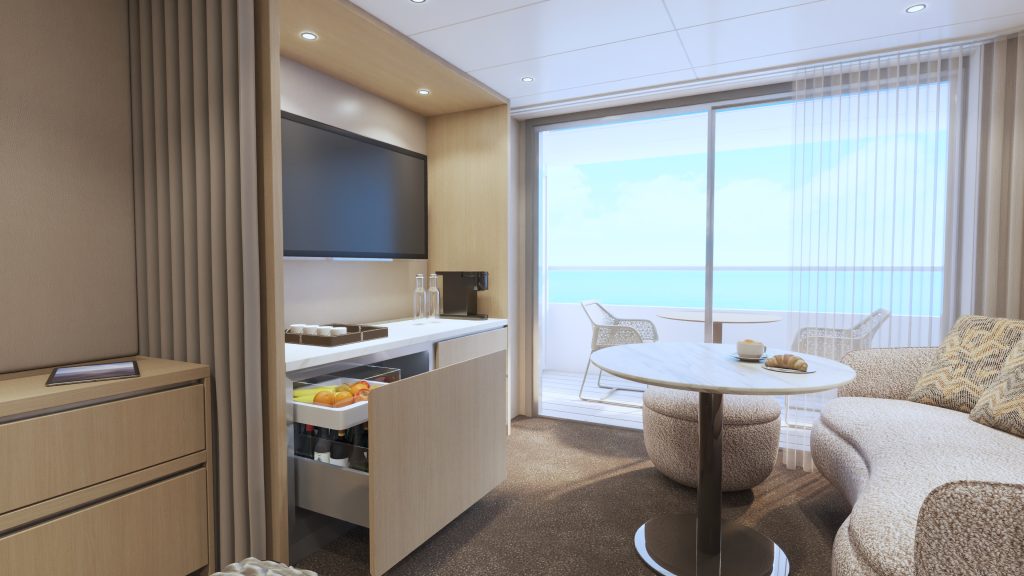 Open-plan hotel suite with a kitchenette, wall-mounted TV, and balcony overlooking the ocean.