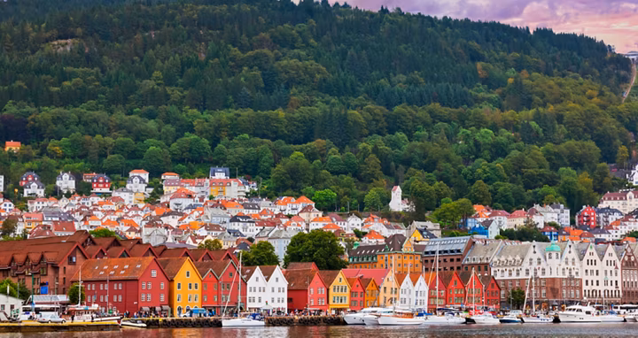 Colorful row of waterfront houses along a calm harbor, with boats and a lush forested hill in the background.