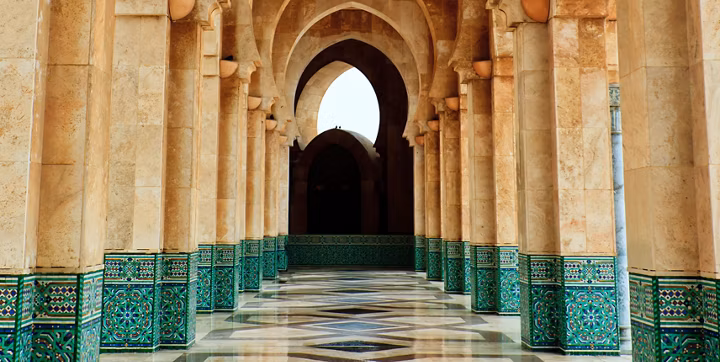 Row of beige stone arches and columns with green tiled bases, converging toward a dark arched doorway at the end.