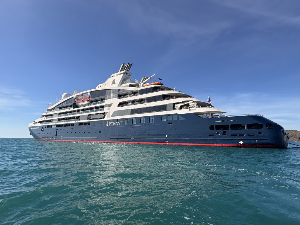 Side view of a large Ponant luxury cruise ship on calm blue water under a clear sky, with lifeboats and multiple decks visible.
