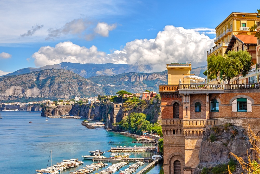 Mediterranean coastal town built on cliffs above a calm blue bay, with terracotta and yellow buildings and a boat-filled marina.