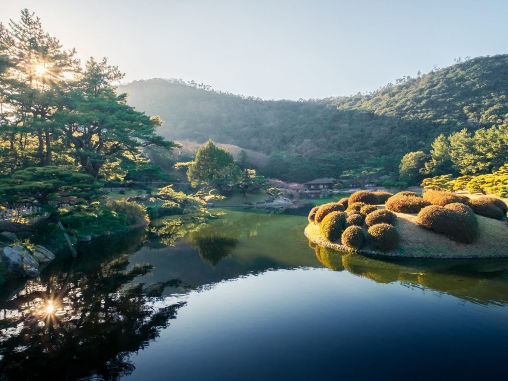 Serene Japanese garden with a reflective pond, sculpted shrubs, and pine trees across a hillside at sunrise or sunset. The sun gleams through trees on the left, mirrored on the water.