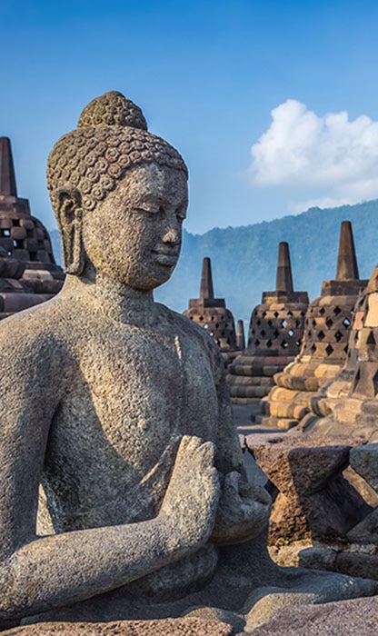 Stone Buddha statue in a meditative pose with surrounding bell-shaped stupas and a clear blue sky at Borobudur Temple.