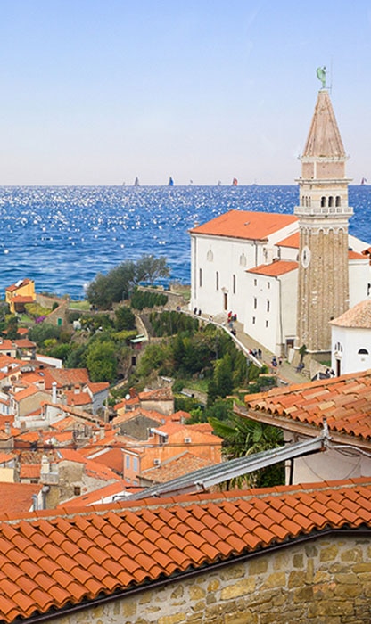 Coastal Mediterranean town with orange-tiled roofs, a tall stone bell tower by a white building, and the blue sea in the background.