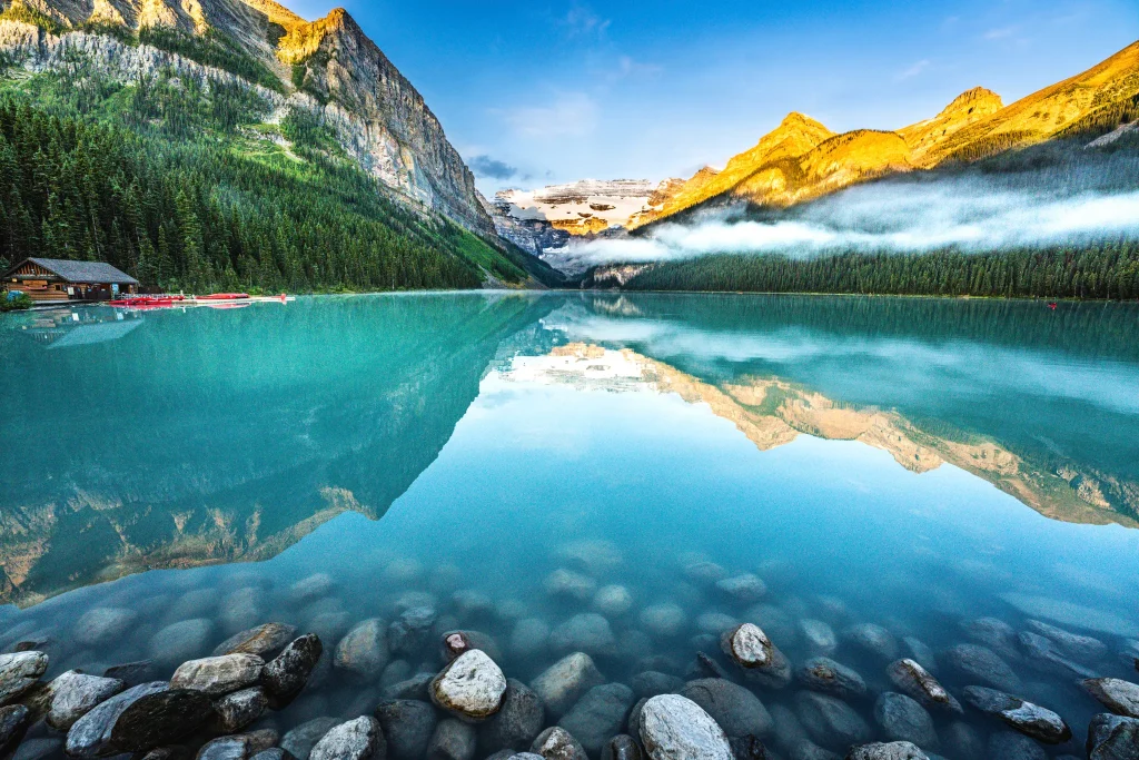Turquoise alpine lake with a rocky shore, dense evergreen forest, and sunlit mountains reflected in calm water at dawns light.