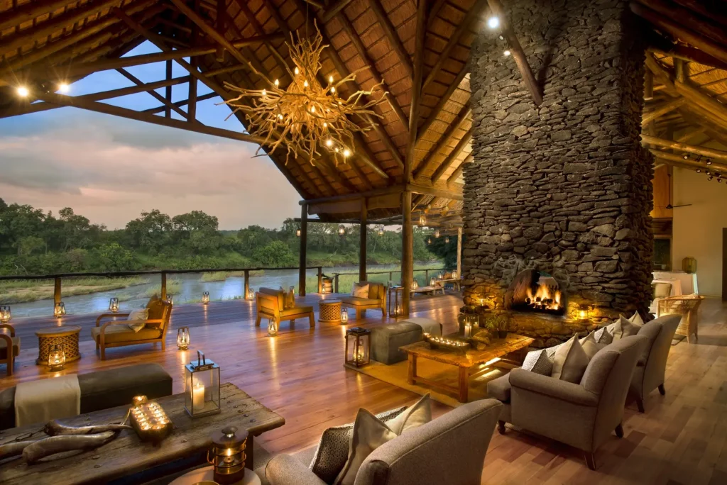 Luxurious riverside lounge with a large stone fireplace, amber lighting, and a wooden deck overlooking a river at dusk.