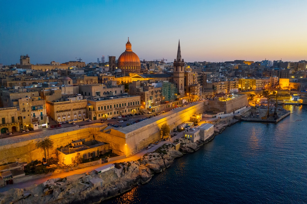 A coastal Mediterranean city skyline at sunset with a domed cathedral and tall church spire overlooking the harbor.