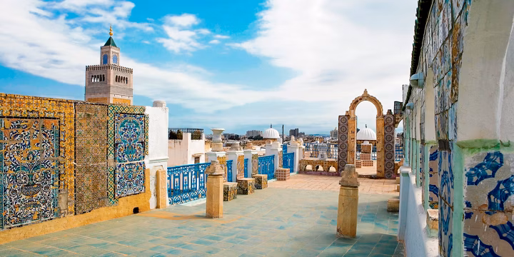 Rooftop terrace with colorful mosaic walls, archway, and turquoise tiles, overlooking a city skyline under a blue sky