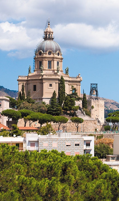 Grand domed church on a hill beside stone fortress, surrounded by trees and a town below.
