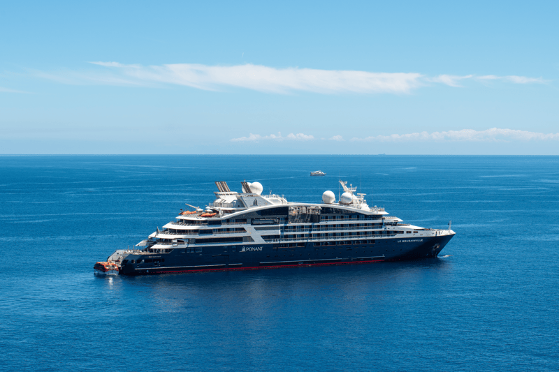 Large luxury cruise ship (Ponant Le Bougainville) sailing in calm blue sea under a clear sky, with distant horizon visible.