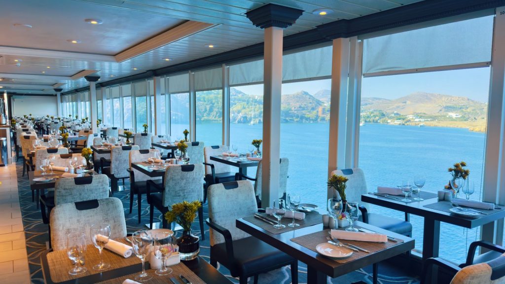 Waterfront dining room with floor-to-ceiling windows, tables set with glassware and yellow flowers, overlooking the sea and hills far away.