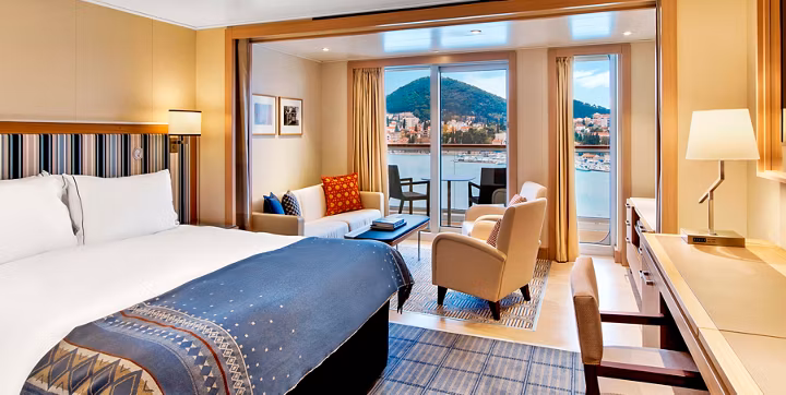 Bright hotel room with a king bed, blue patterned bedspread, seating area, and a balcony with a lake and mountain view