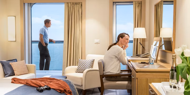 In a bright hotel suite, a woman sits at a wooden desk with a mirror, smiling as she talks on the phone; a man stands on the balcony looking out at the sea.