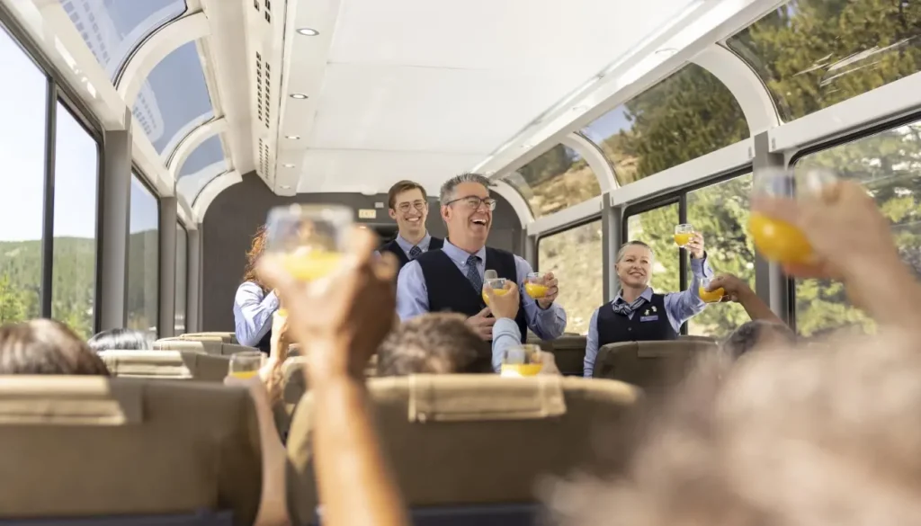 Coworkers toasting with orange juice inside a bright sightseeing coach or train car during a celebration gesture.