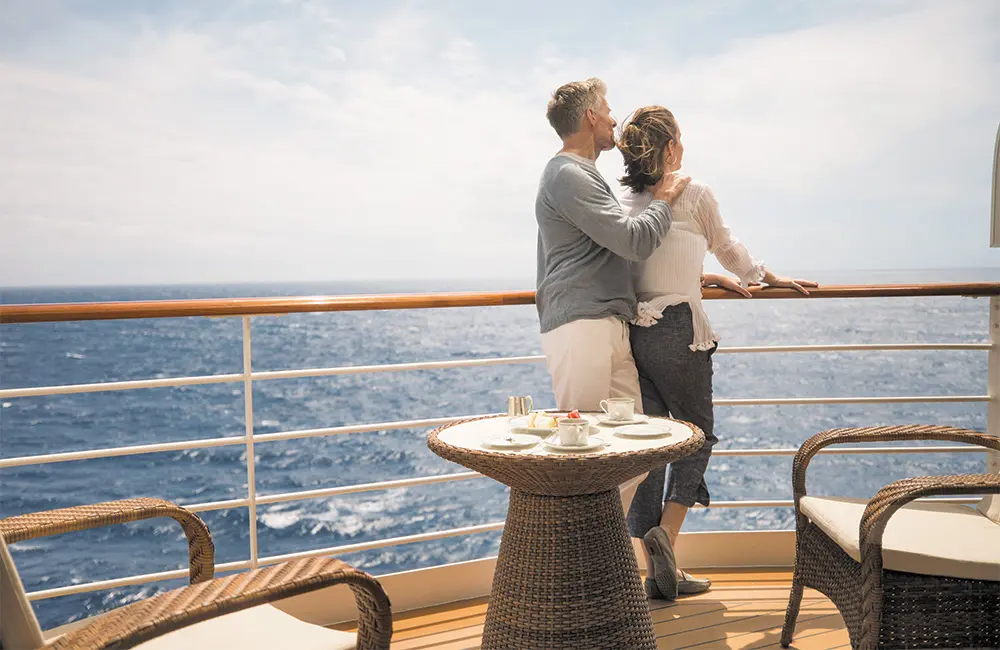 Couple standing at a cruise ship balcony, embracing and gazing at the ocean with a small tea setup on a wicker table nearby.