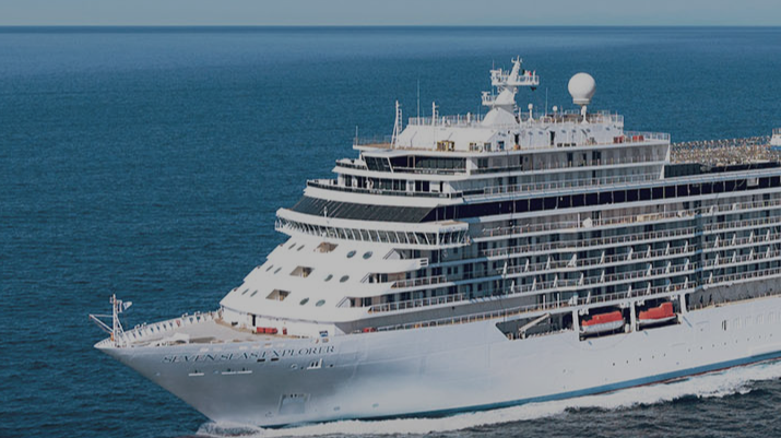Large white cruise ship sailing on calm blue sea with multiple decks visible and red lifeboats along the side, viewed from bow-left angle.