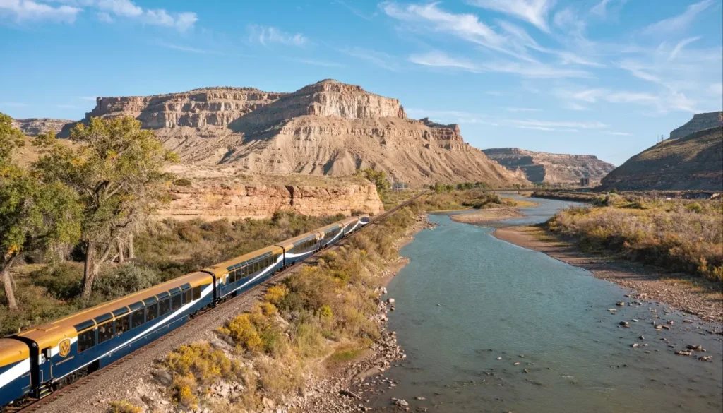 Passenger train traveling along a river through a desert canyon under a blue sky.