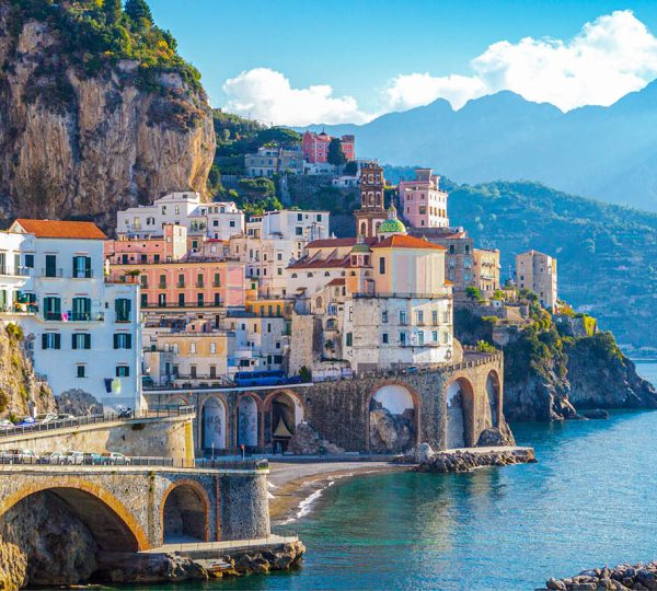Colorful houses cling to a cliff above clear blue water in a coastal Italian town.