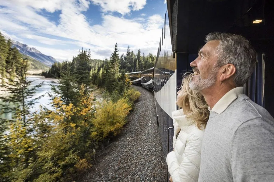 Couple leaning from a train window, smiling as they watch a forested river valley roll by.